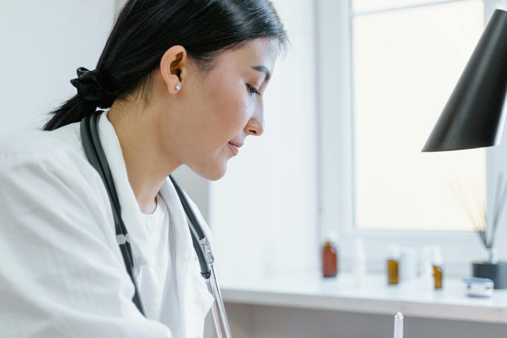 A woman in a white coat with a stethoscope around her neck is looking down, possibly writing or reading. She has dark hair pulled back and is in a bright room with a window and blurred bottles in the background.