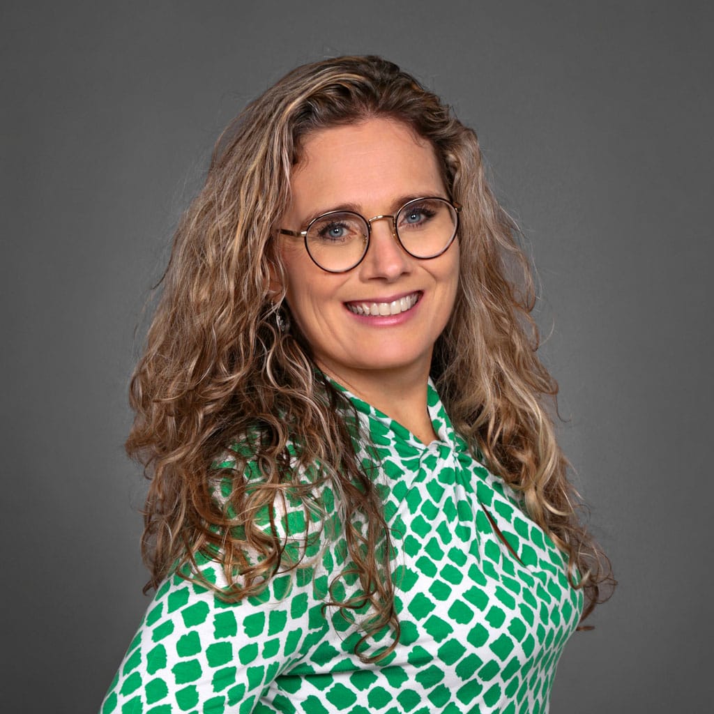 A woman with long, curly blonde hair smiles at the camera. She wears round glasses and a green and white patterned blouse with long sleeves. The background is plain gray, and she stands at an angle with her hand on her hip.