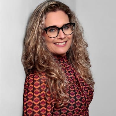 A woman with long, curly blonde hair and black-rimmed glasses smiles at the camera. She is wearing a high-neck, long-sleeved top with a geometric red, pink, and orange pattern, and is standing against a plain light gray background.