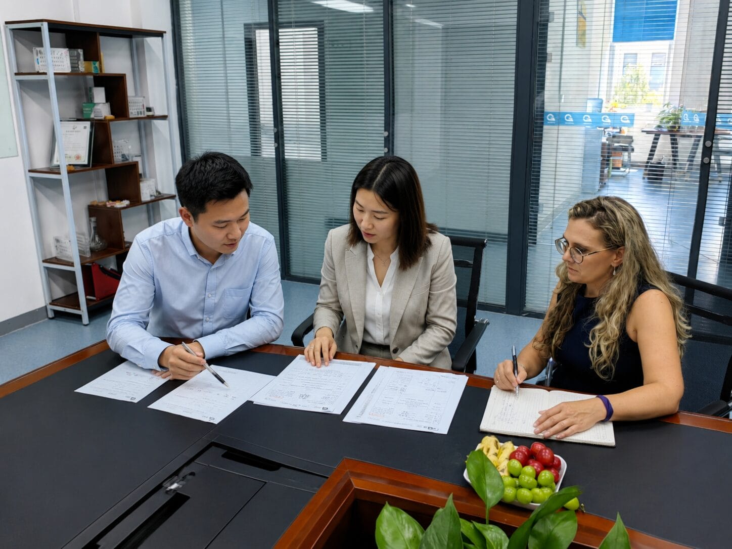 Three people sit at a conference table reviewing documents. The two on the left wear business attire; the one on the right has glasses and long, wavy hair, taking notes. Papers and fruit plates are on the table, with plants and office shelves in the background.